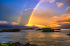 A vibrant double rainbow stretching across a dramatic, glowing golden sunset sky over a calm ocean bay with dark rocky islands.
